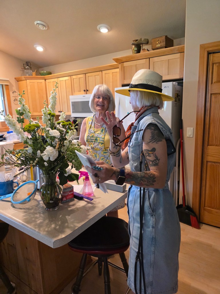 Two women engaging in conversation in a kitchen, one wearing a straw hat and denim dress, and the other in a colorful outfit, with a vase of flowers on the table.