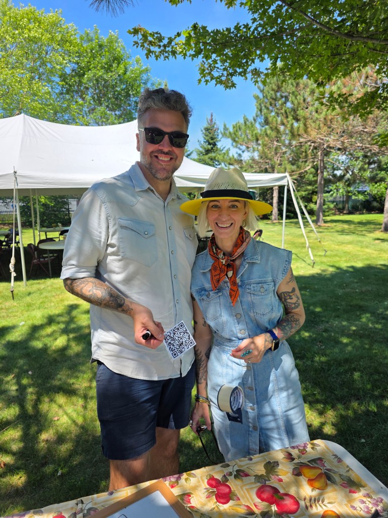 A man and a woman smiling outdoors at a summer event, holding a QR code. The man wears sunglasses and a light shirt, and the woman wears a denim dress with an orange scarf. In the background, there are trees and a tent.