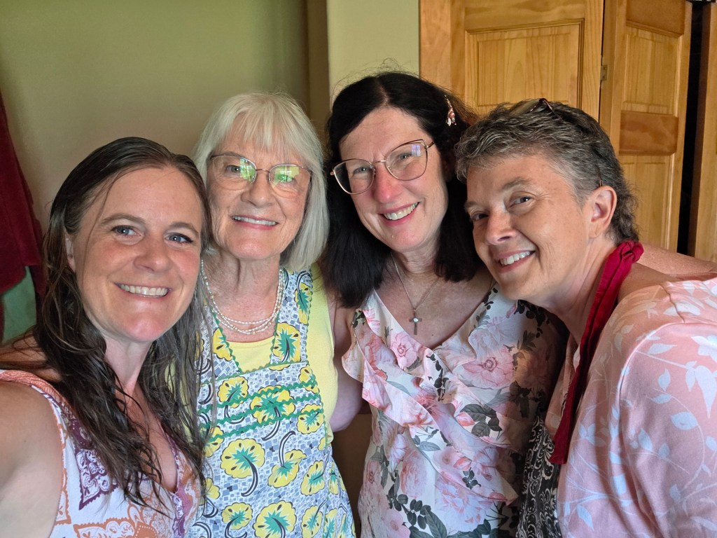 A group of four women smiling together in a cozy indoor setting, dressed in colorful outfits and aprons.