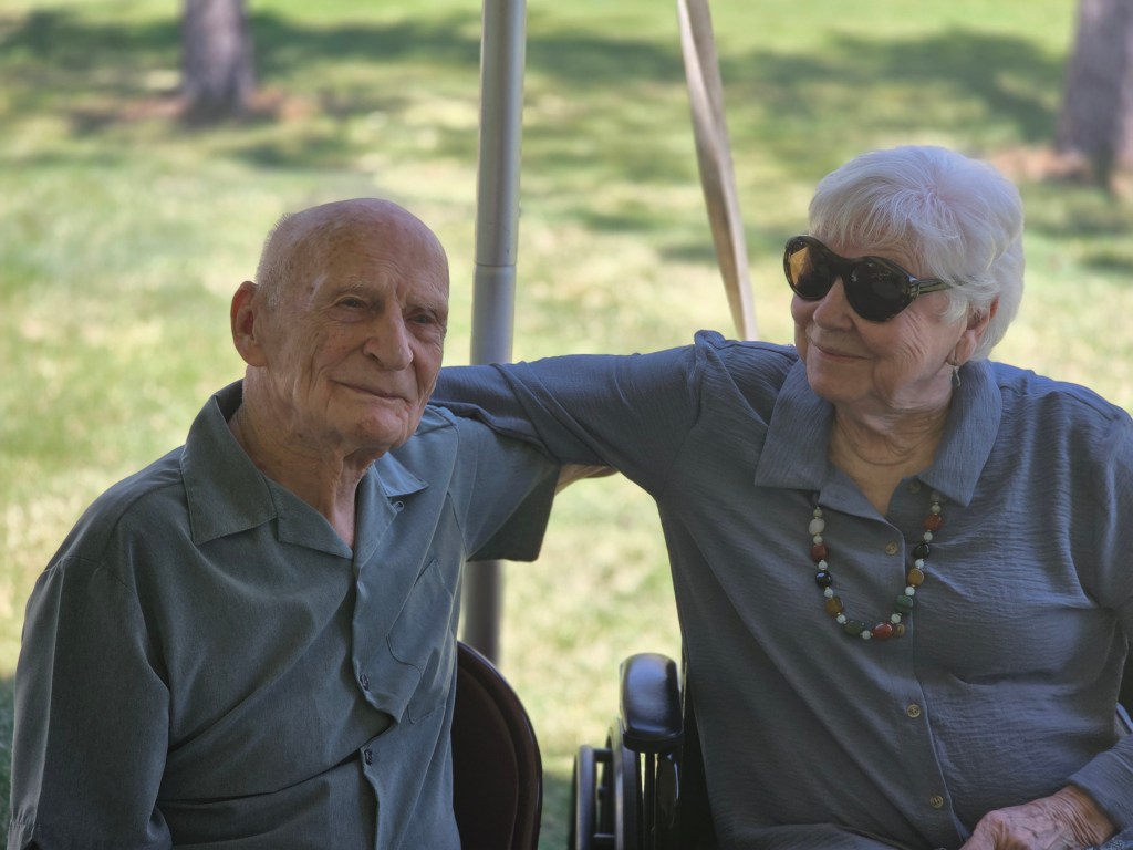 An elderly man wearing a blue shirt and a woman in a blue blouse with sunglasses, sitting together outdoors under a tent.