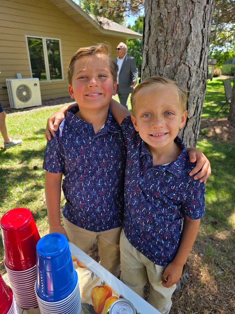 Two smiling boys standing closely together outdoors, both wearing matching navy blue floral shirts and light-colored pants, with a table of colorful cups and refreshments in the foreground.
