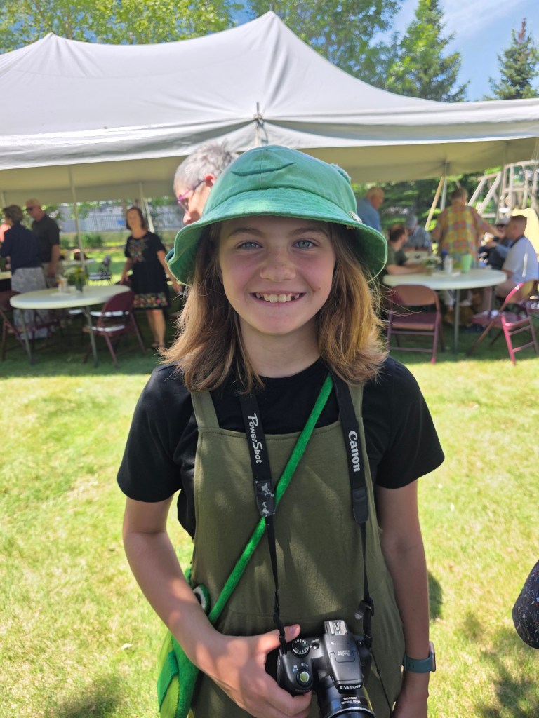 A smiling young person wearing a green bucket hat and an olive green apron over a black shirt, holding a Canon camera, standing in a grassy area with people in the background.