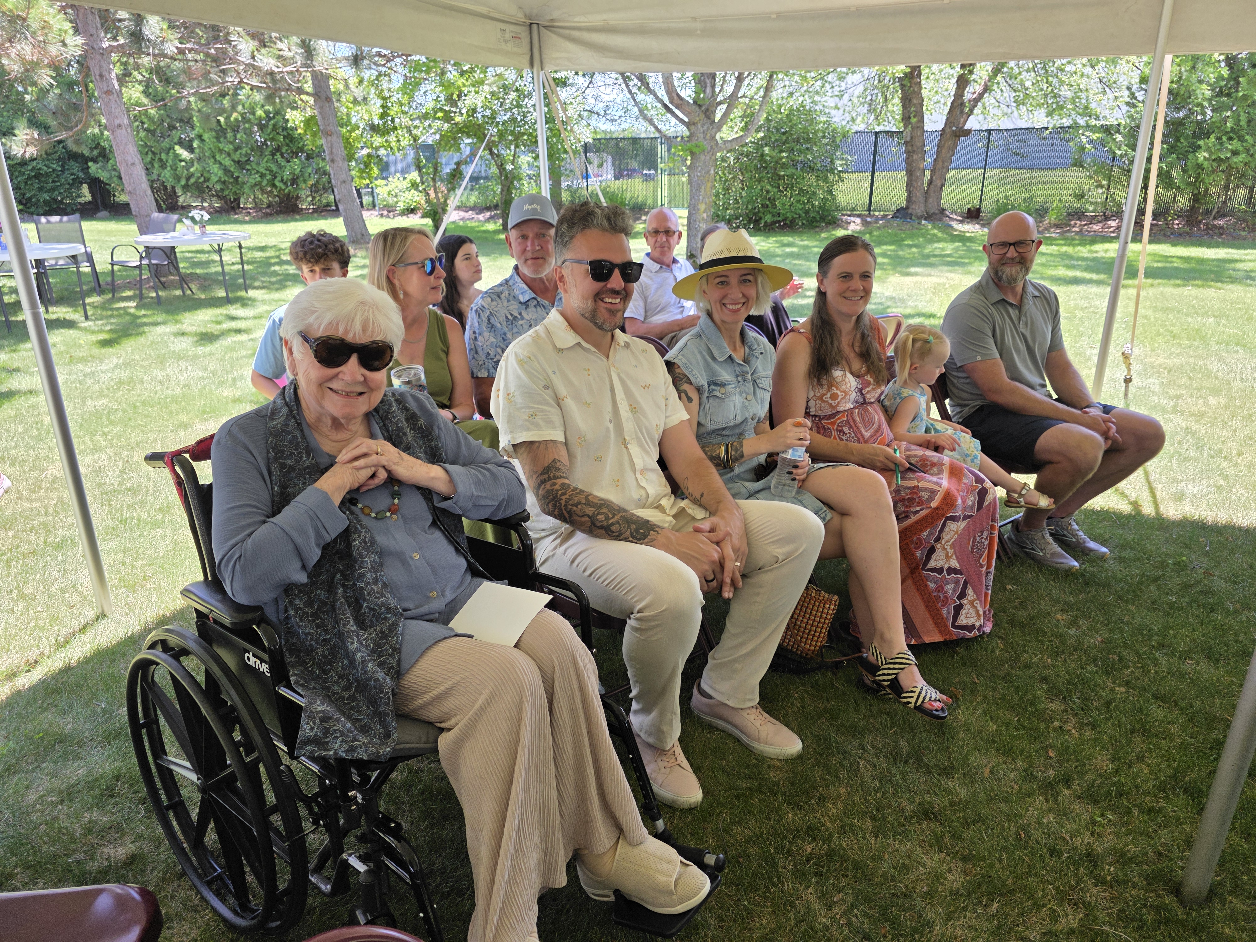 A diverse group of people sitting in a shaded outdoor setting under a tent, with green grass and trees in the background. Front row includes an elderly woman in a wheelchair wearing sunglasses, and a couple of younger adults, along with others enjoying the sunny day.