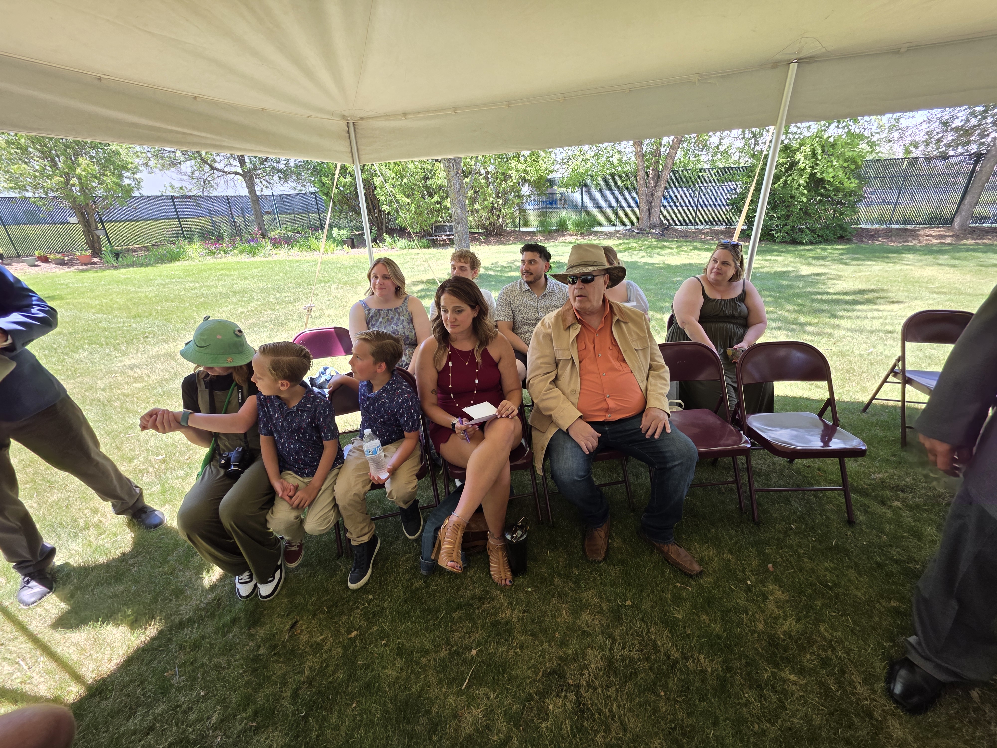 A group of adults and children sitting under a tent at an outdoor event. Some children are wearing matching shirts, while adults are casually dressed. The background features green grass and trees.
