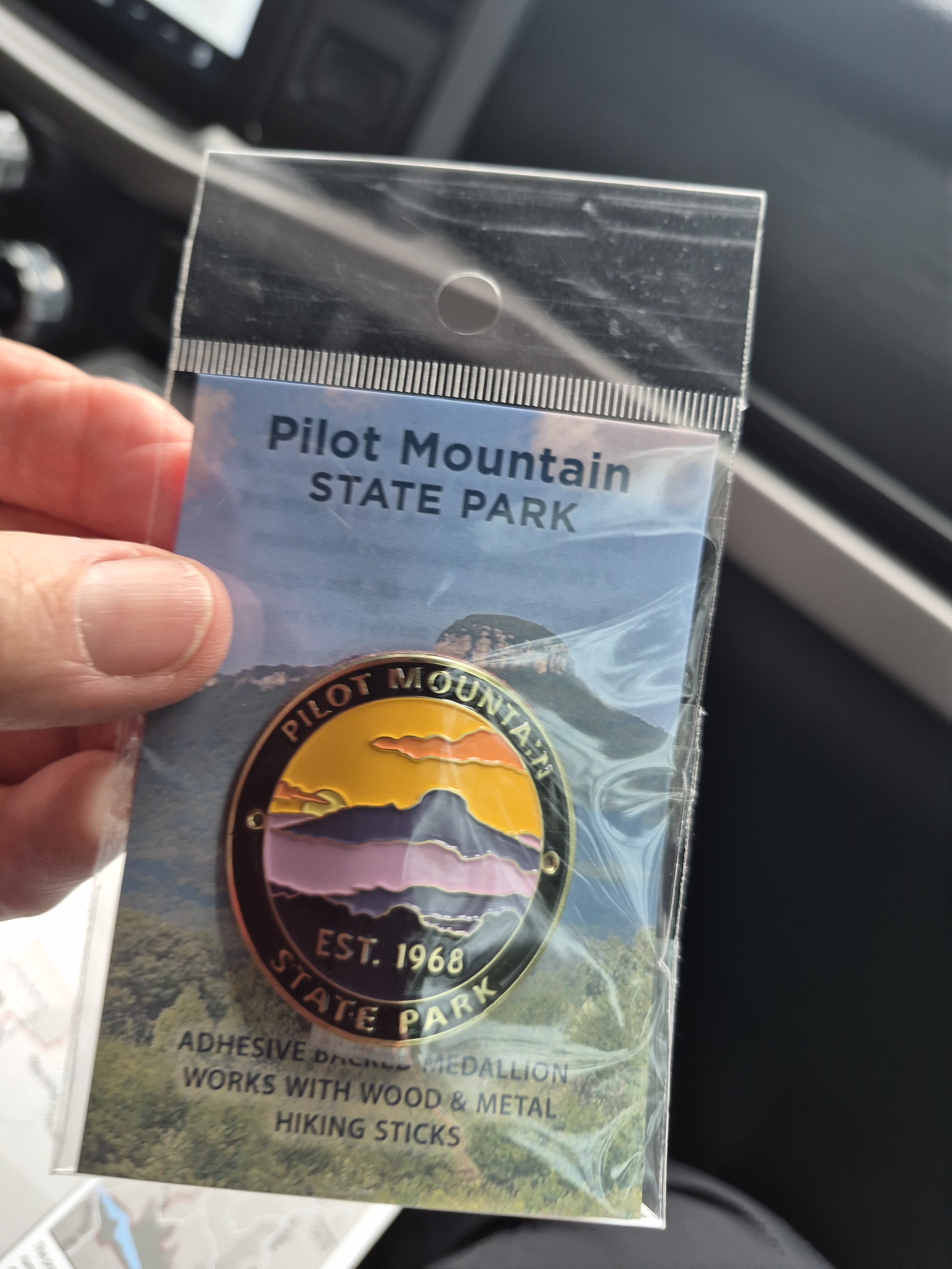 A hand holding a packaged adhesive medallion featuring Pilot Mountain State Park, showcasing mountains and a sunset graphic, with text indicating its suitability for wood and metal hiking sticks.