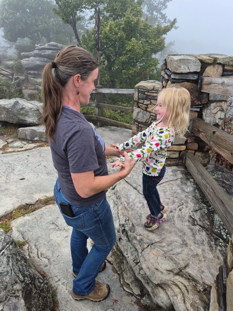 A woman and a young girl playfully hold hands on a rocky outdoor setting, surrounded by fog and trees.