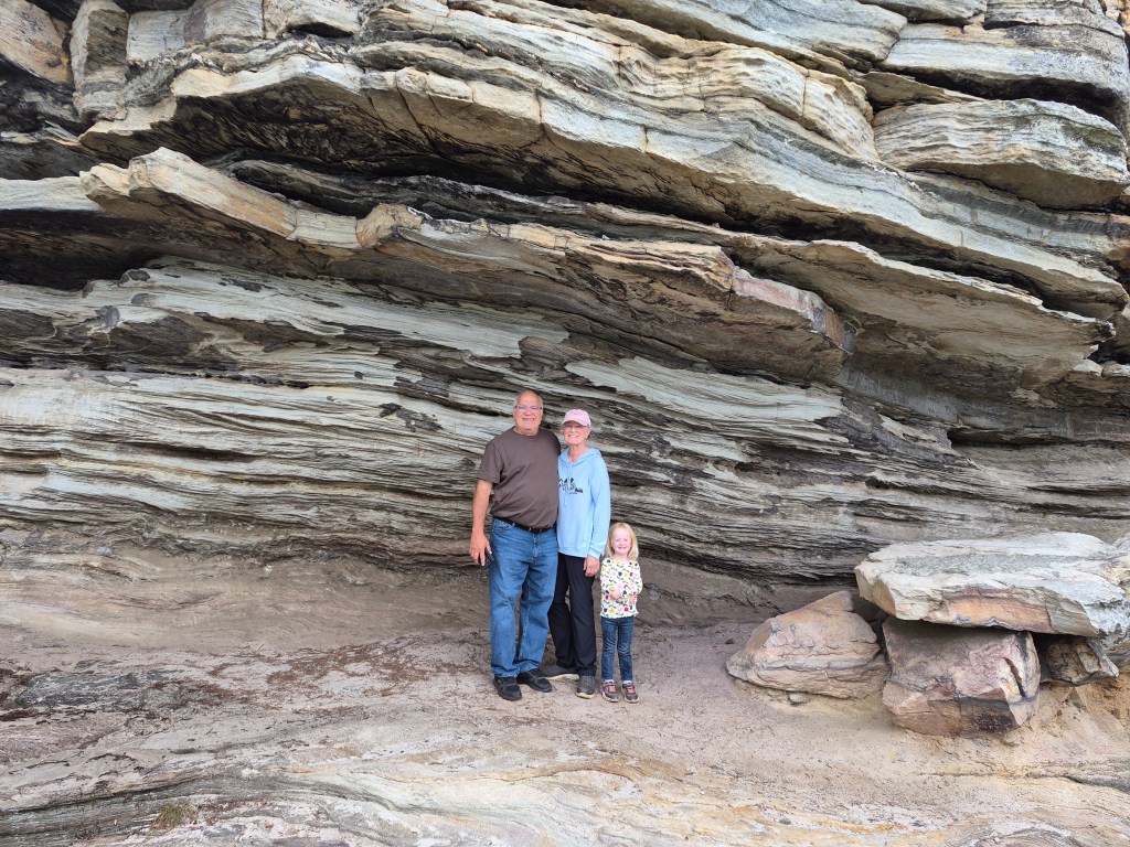 A family stands together at a rocky formation, featuring layered stone structures. The group includes an adult man, an adult woman, and a young girl, all smiling and enjoying the outdoors.