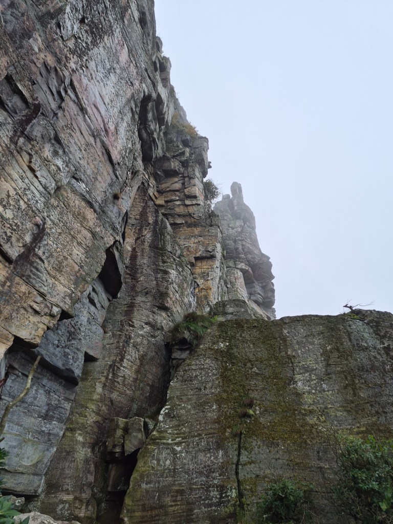 A steep rock formation rising into a foggy sky, featuring rugged textures and moss-covered surfaces.