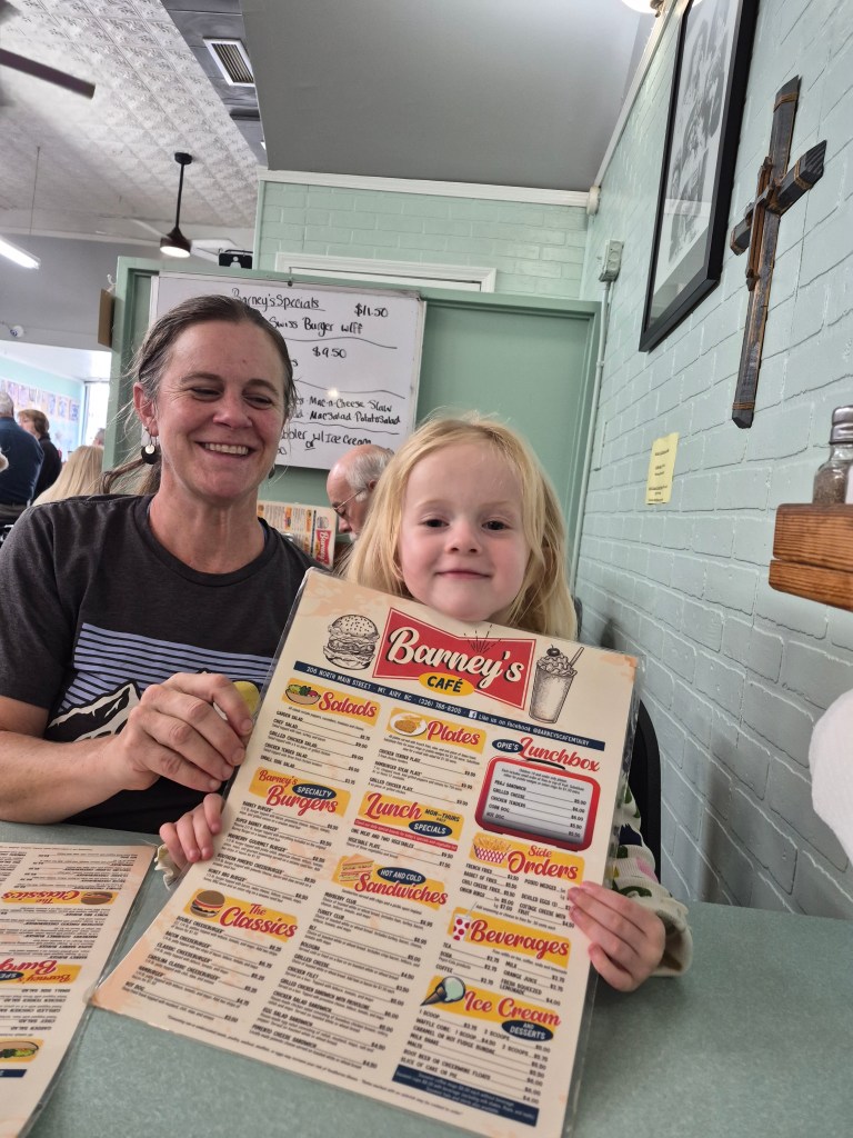 A woman and a young girl smiling while holding a menu at Barney's Café, showcasing sections for salads, burgers, lunches, sandwiches, beverages, and ice cream.
