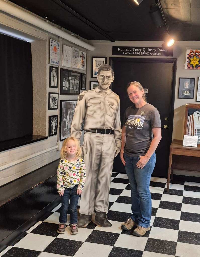 A smiling woman and a young girl pose next to a life-sized cardboard cutout of a man in police uniform in a museum setting, showcasing a black and white checkered floor and framed photographs on the wall.