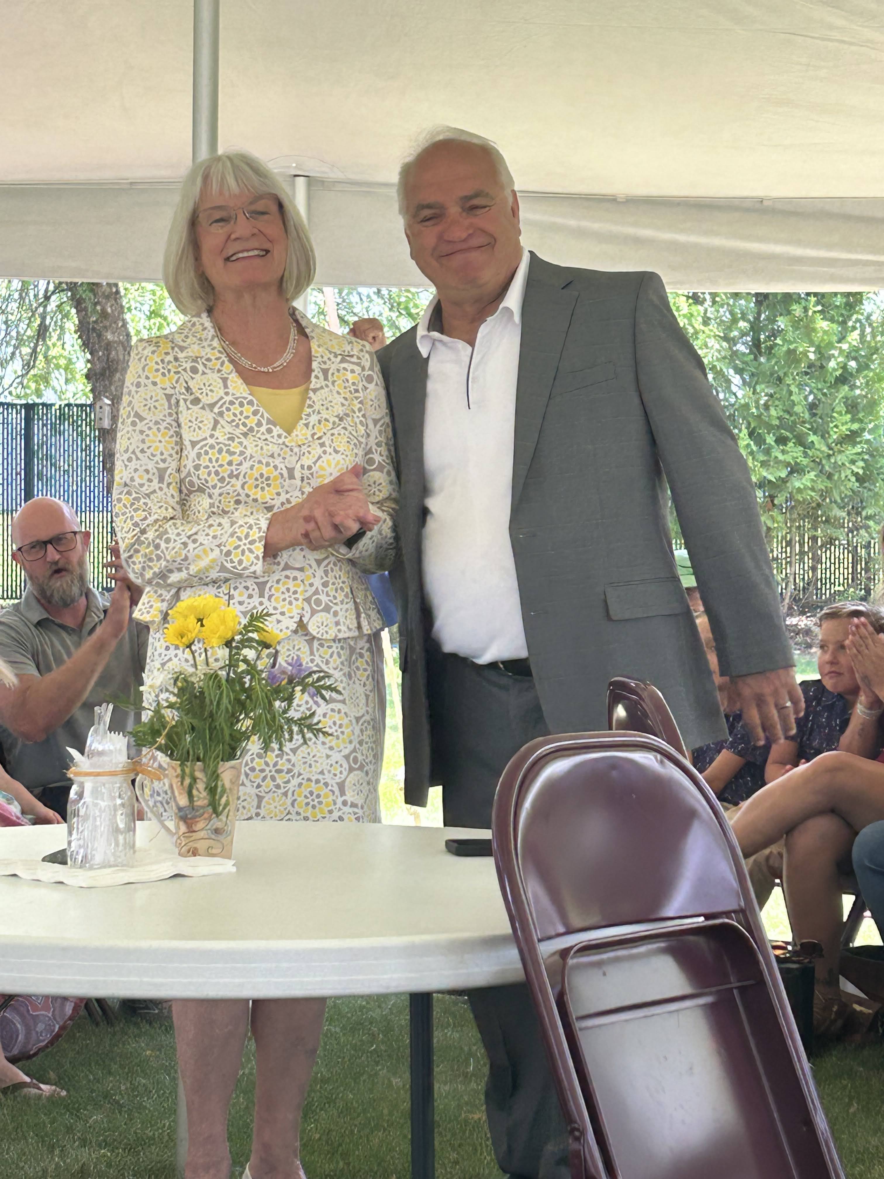 A smiling woman in a floral-patterned suit and a man in a gray suit stand together, holding hands under a tent. A table with a flower vase is visible in the foreground, and guests can be seen seated in the background.