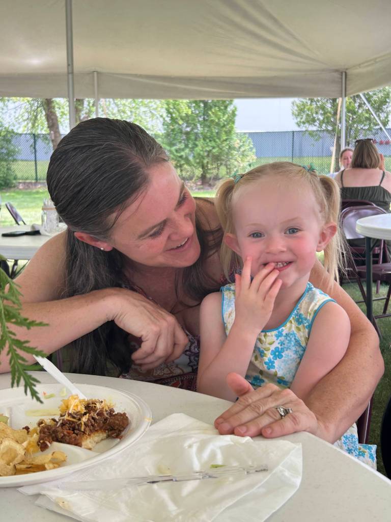 A smiling woman and a young girl sitting at a table under a tent, enjoying a meal together. The girl is playfully covering her mouth while the woman looks at her affectionately. In the foreground, there is a plate of food and some napkins.