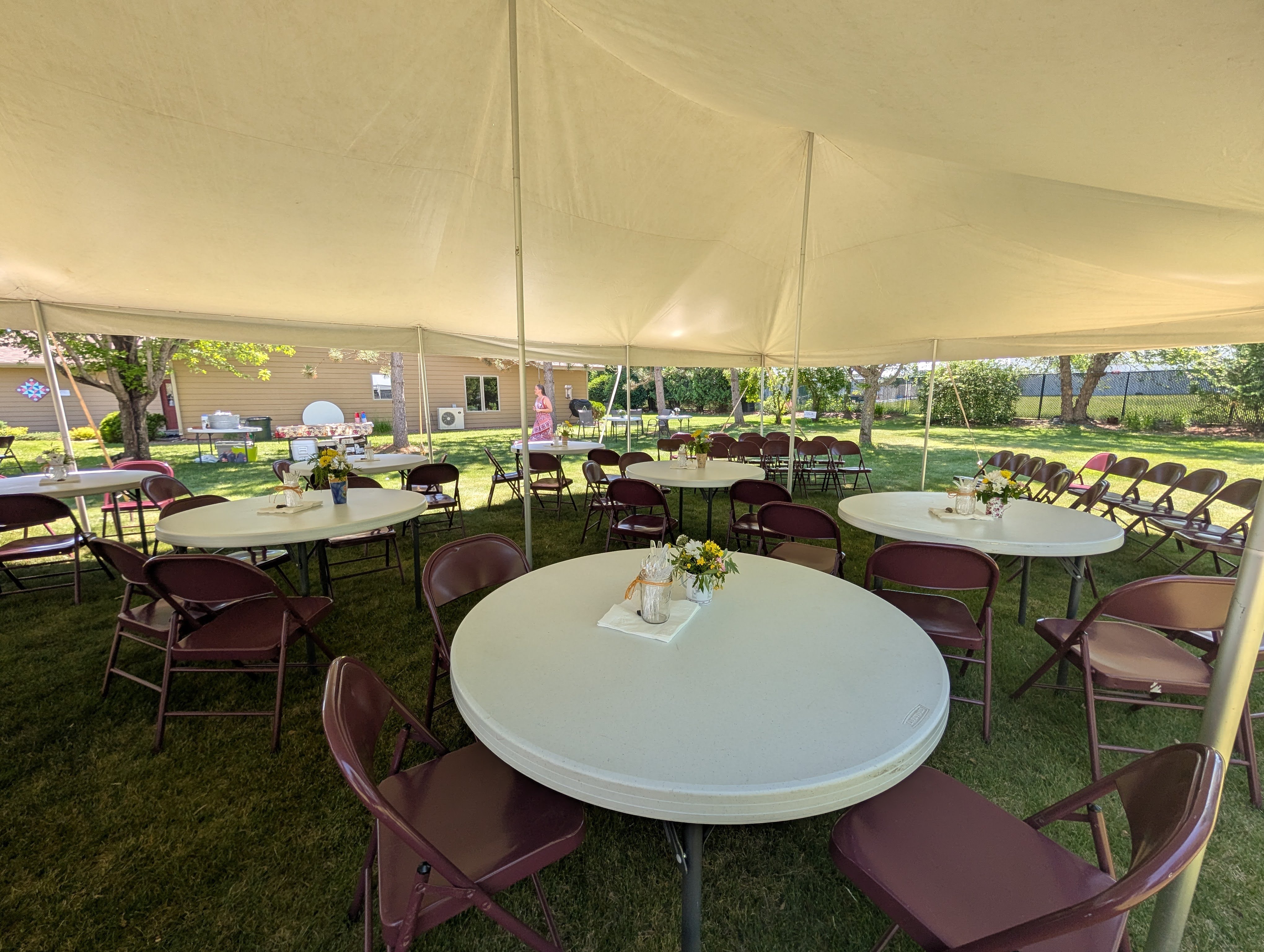A large outdoor tent filled with round tables and chairs set up for an event, featuring small flower arrangements on the tables and a view of the grassy area outside.