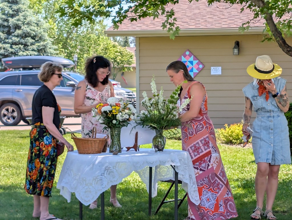A group of four women arranging flowers at a table outdoors, with greenery and a house in the background.