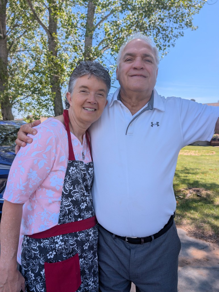 A smiling middle-aged couple posing together outdoors, with trees in the background. The woman wears a floral shirt and an apron, while the man is dressed in a polo shirt and gray pants.