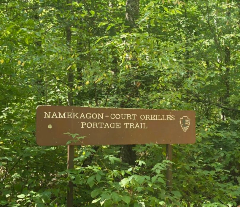 A sign reading 'NAMEKAGON-COURT OREILLES PORTAGE TRAIL' surrounded by lush green foliage.