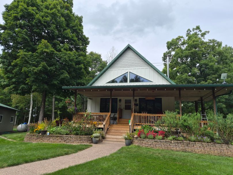 A modern cabin with a triangular roof, featuring a large porch with wooden steps, surrounded by colorful flowers and greenery.