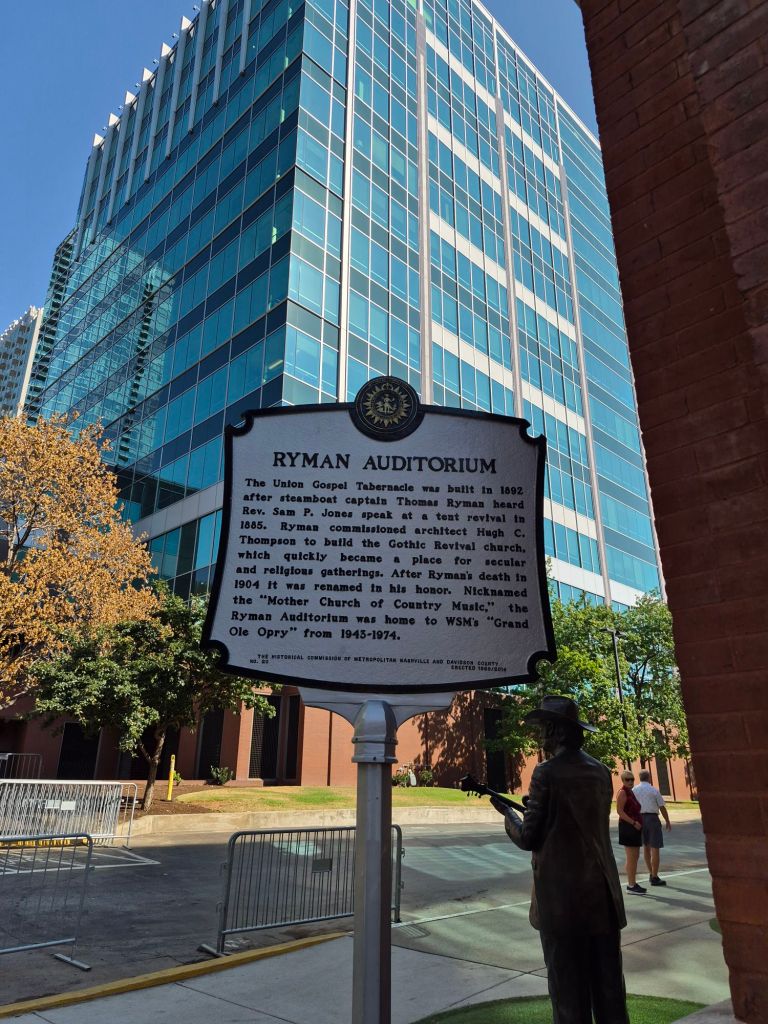 A historical marker for the Ryman Auditorium, detailing its background and significance, against a backdrop of modern buildings. A statue of a man in a suit stands beside the marker.
