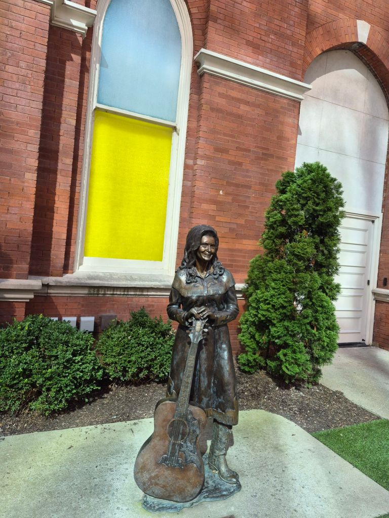 A bronze statue of a woman holding a guitar, standing in front of a brick building with colorful windows and greenery surrounding the base.