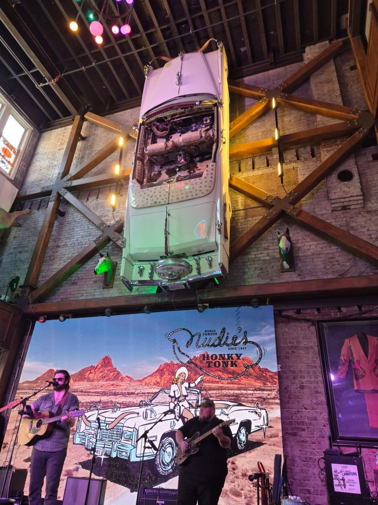 A colorful interior of a honky-tonk with a vintage car suspended from the ceiling, musicians performing on stage in the foreground, and a large mural of a desert landscape in the background.