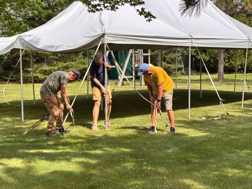 Three men setting up a tent on a grassy area, each using a pole to secure the structure.